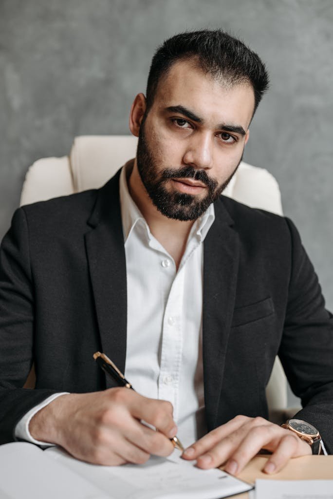 Serious man in a black suit signing documents in an office setting.